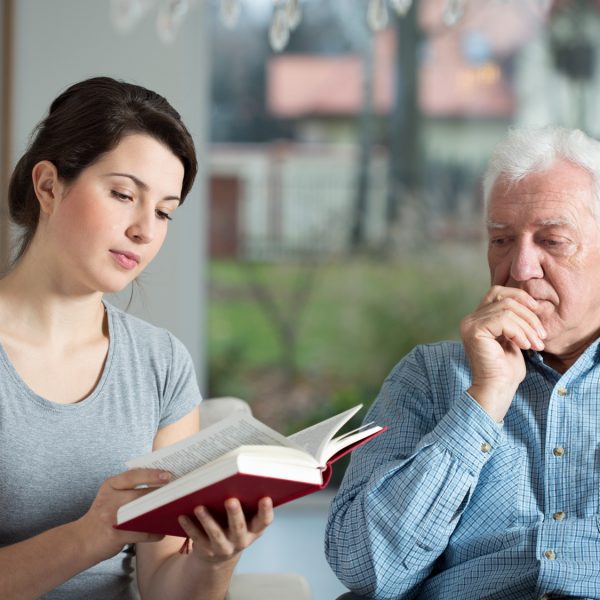 Young attractive caregiver reading the book to aged man